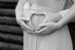 A loving couple forms a heart shape over a pregnant belly in a black and white portrait.
